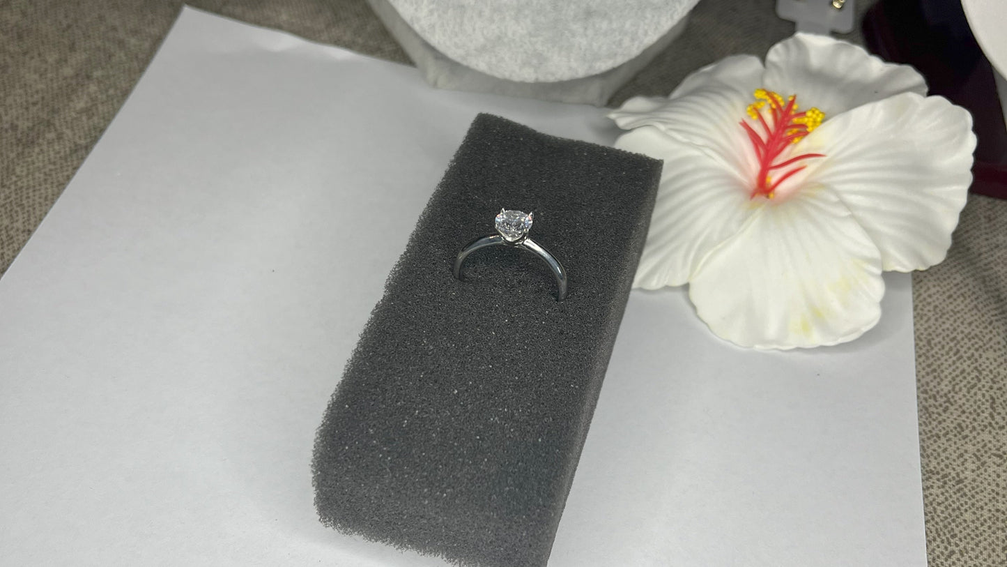 Anello solitario in acciaio placcato argento con pietra brillante centrale, montatura classica elegante su superficie grigia con conchiglia decorativa
