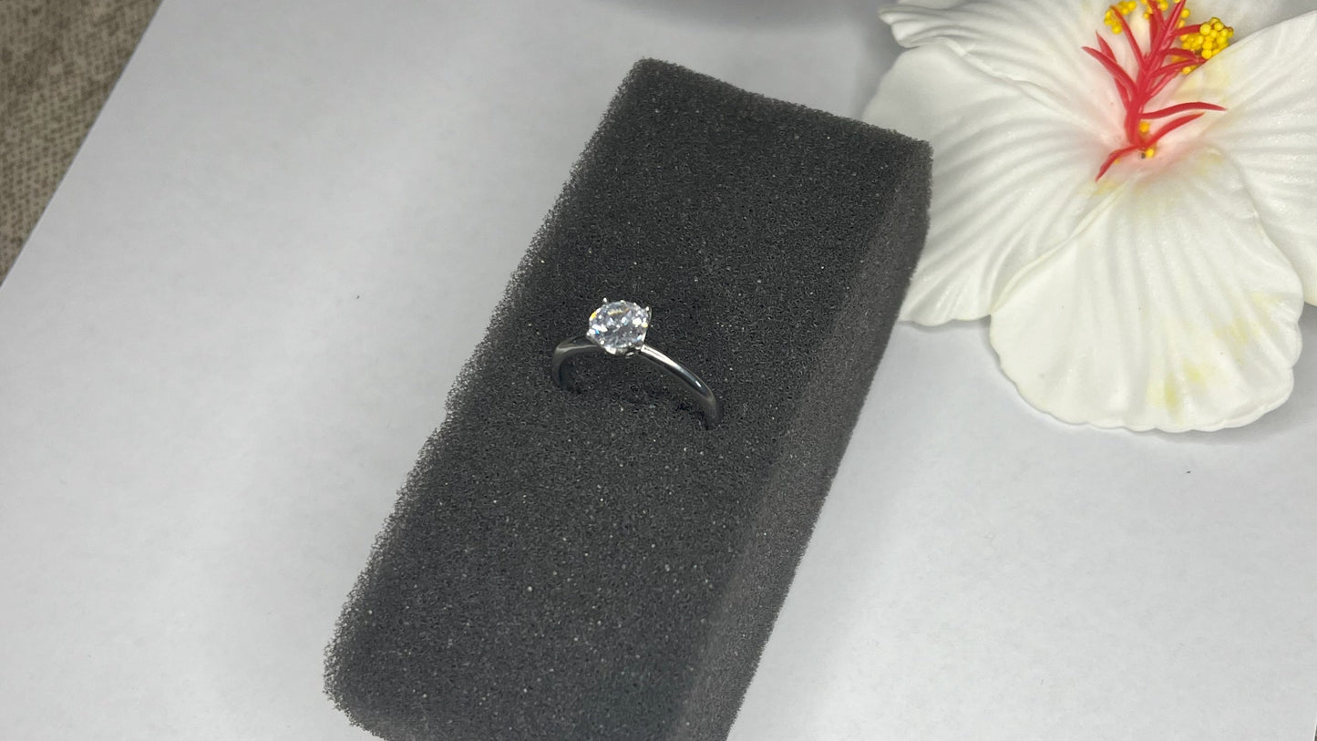 Anello solitario in acciaio placcato argento con pietra brillante centrale, montatura classica elegante su superficie grigia con conchiglia decorativa