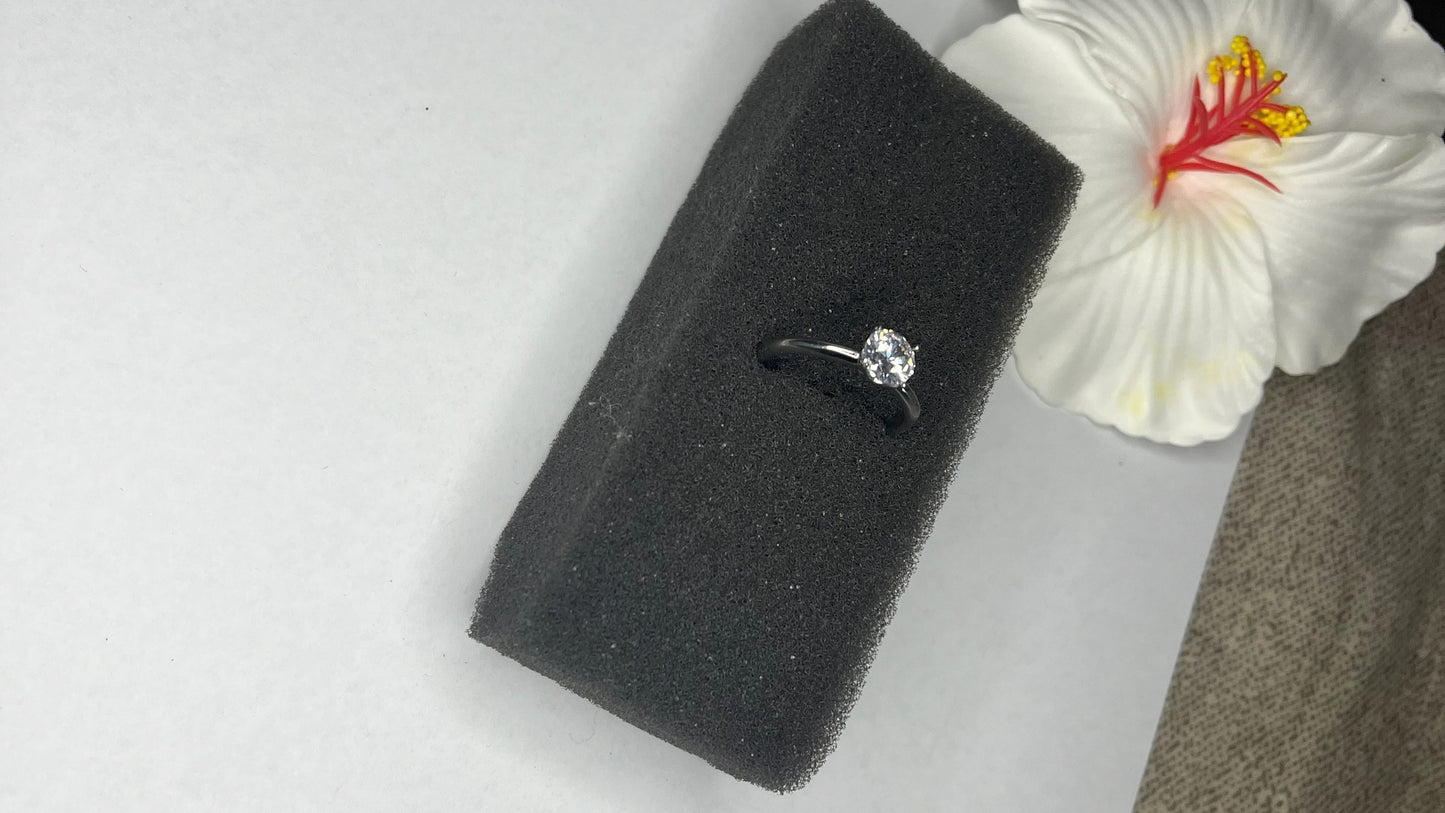 Anello solitario in acciaio placcato argento con pietra brillante centrale, montatura classica elegante su superficie grigia con conchiglia decorativa
