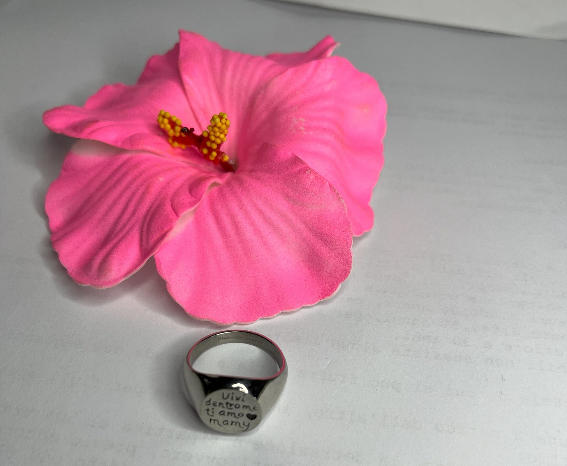 Anello Regina Mamy in acciaio premium placcato argento con incisione dedicata alla maternità, fotografato su superficie bianca con fiore rosa