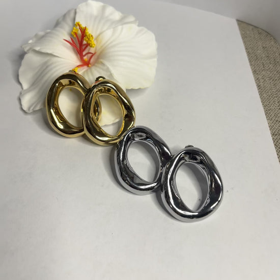 Orecchini Wave premium in due varianti - placcati oro e argento con design organico fluido a forma di onda, fotografati accanto a fiore bianco