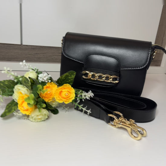 Borsa Toscana Chic in vera pelle nera Made in Italy con catena dorata decorativa e tracolla regolabile, fotografata su superficie bianca con fiori gialli