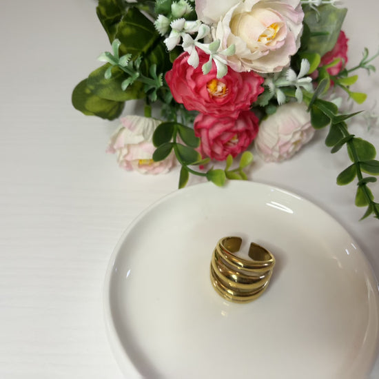 Anello Fascia Tripla placcato oro con tre fasce sovrapposte eleganti, design moderno e minimalista su piattino bianco con fiore rosa e bianco 