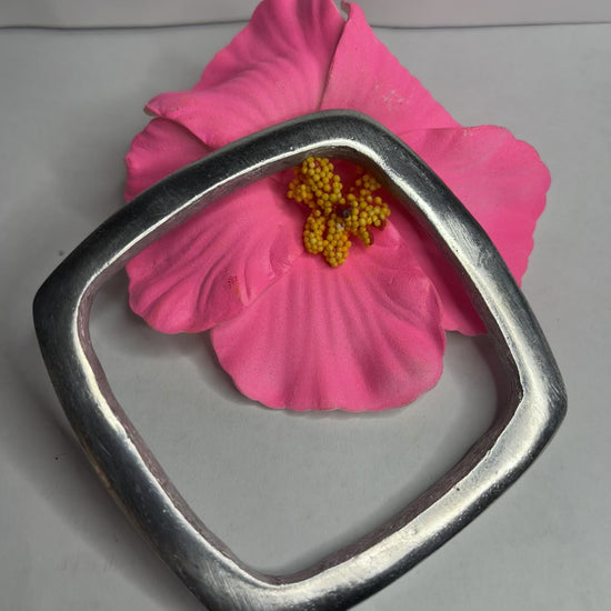 Bracciale quadro in alluminio con design geometrico moderno e finitura satinata, fotografato accanto a fiore rosa