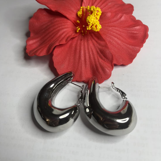Orecchini Della Schiava a cerchio in acciaio premium placcato argento con superficie liscia e brillante, chiusura a scatto sicura, fotografati su superficie bianca e fiore rosso