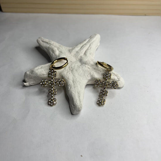 Orecchini pendenti con ciondoli a croce decorati con cristalli diamantati e ganci dorati, appoggiati su stella marina bianca