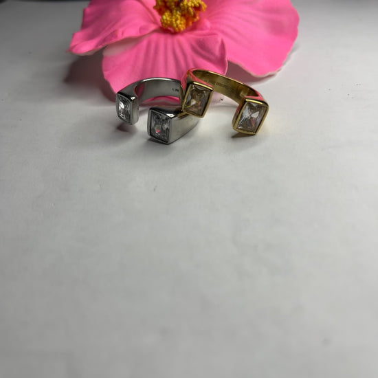Anello Geometric Luxury con design geometrico quadrato, disponibile in versione placcata oro e argento, esposto su fiore rosa decorativo