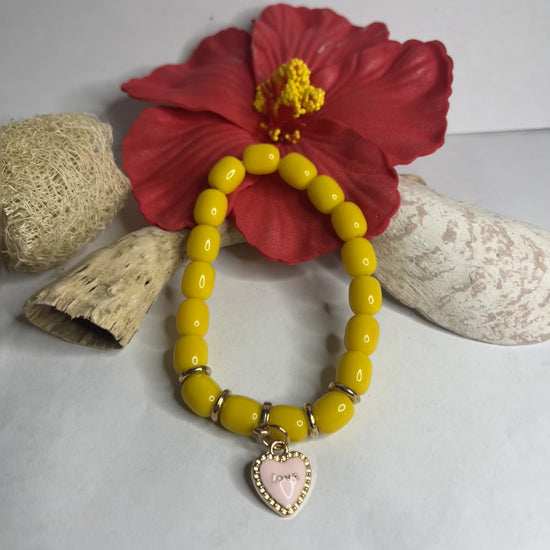 Braccialetto elastico con perle gialle dorate e ciondolo a cuore rosa su sfondo neutro