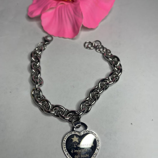 Braccialetto chunky argentato con pendente a cuore nero contenente la scritta "L'essenziale è invisibile agli occhi" su sfondo grigio con petali rosa