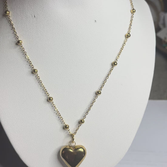  Collana Allegra in acciaio premium con catena decorata da sfere e ciondolo cuore, variante placcato oro, esposta su busto espositore bianco 